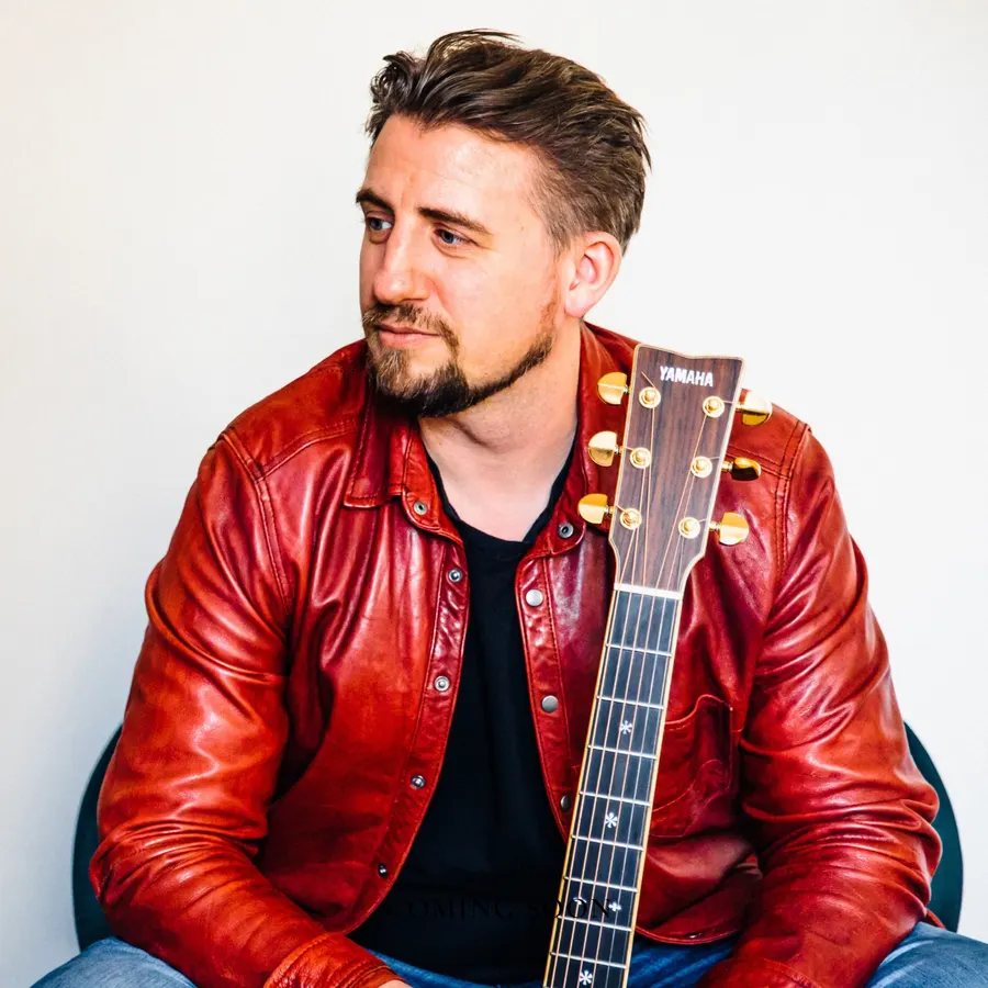 Ryan Inglis in a red leather jacket holding his guitar